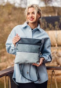 Graue und marineblaue Leder-Crossbody-Tasche mit Reißverschluss, die ein geteiltes Design und einen verstellbaren Träger aufweist, vor einem natürlichen Hintergrund gehalten.