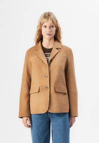 Kvinne iført en tan semsket blazer over en mønstret genser og blå jeans, stående mot en ensfarget hvit bakgrunn.