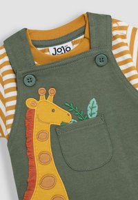 Salopette en coton verte avec un design de poche brodée de girafe, accompagnée d'une chemise rayée moutarde et blanche en dessous.