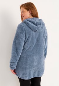 Flauschige blaue Kapuzenjacke aus weichem Material, mit langen Ärmeln und abgerundetem Saum, von hinten gesehen.