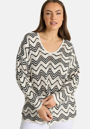 Vrouw met lang bruin haar, gekleed in een wit gebreid vest met zwarte zigzagpatronen, staand tegen een effen lichte achtergrond.