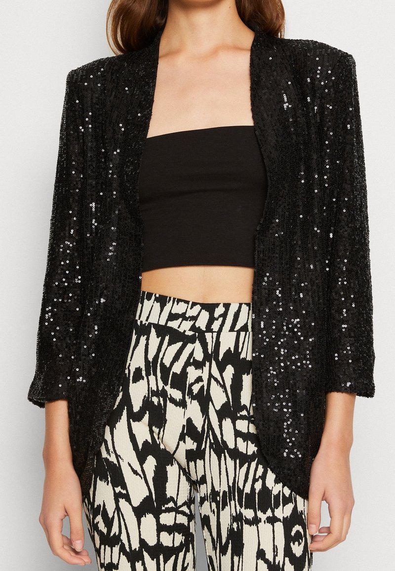 Femme portant une veste noire ouverte à sequins, un crop top noir sans bretelles, et un pantalon taille haute noir et blanc à motifs.