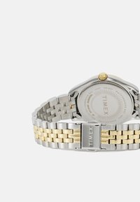 Montre-bracelet en acier inoxydable argent et or avec un boîtier rond, un bracelet en maille et un cadran blanc texturé. Dotée d'une couronne et d'un fermoir jaunes.