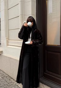 Naine musta karvkatte ja pearätikuga joob valgest tassist, hoides alustassi, seisab munakiviteel hoone nurgal.