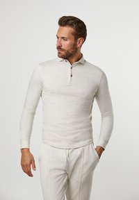 Light beige long-sleeve polo shirt with a ribbed collar, three-button placket, and soft, textured fabric. Worn with matching pants.