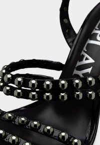Μαύρα πέδιλα με τακούνι από δερμάτινη βερνικωμένη επιφάνεια, με πολλαπλούς λεπτούς ιμάντες διακοσμημένους με στρογγυλά μεταλλικά τρουκς και λεία μαύρη σόλα.