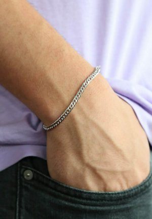 Silver metal chain bracelet on a wrist, featuring interlocking links with a smooth texture, complementing a light purple shirt and dark jeans.
