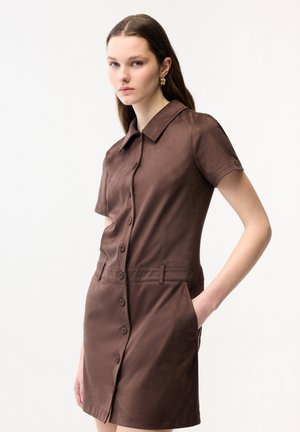 Giovane donna che indossa un vestito marrone a maniche corte con bottoni e tasche laterali, in piedi con una mano in tasca davanti a uno sfondo semplice.