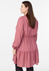 Mujer con largo cabello castaño, vistiendo un vestido de manga larga en un tono rosa empolvado con textura, de pie de perfil contra un fondo blanco.