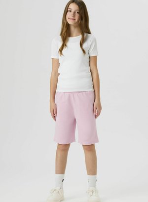 Adolescente debout, portant une chemise blanche à manches courtes, un short rose pâle jusqu'aux genoux, des chaussettes blanches et des baskets blanches, fond neutre.