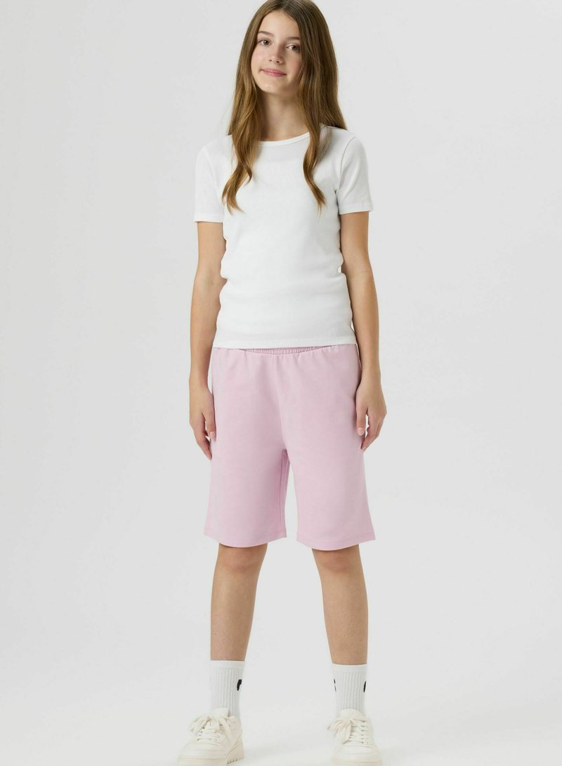 Adolescente debout, portant une chemise blanche à manches courtes, un short rose pâle jusqu'aux genoux, des chaussettes blanches et des baskets blanches, fond neutre.