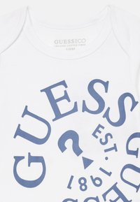 Biely organický bavlnený overal s výraznou modrou typografiou, vrátane textu "GUESS," "EST. 1981" a otázníka, usporiadaný v dynamickom rozložení.