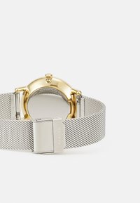 Stříbrný síťkový náramek hodinek s kulatým pouzdrem z nerezové oceli v zlatém odstínu. Obsahuje hladký, lesklý povrch a sponu s logem.