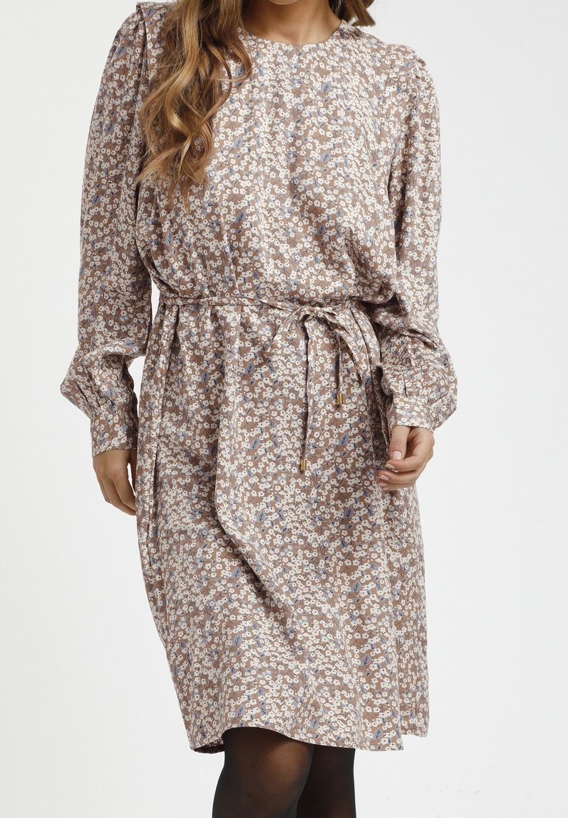 Femme portant une robe beige à manches longues, longueur genoux, avec un petit motif floral blanc et bleu et une ceinture à nouer à la taille.