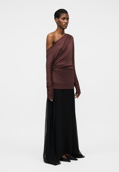 Kobieta stoi, ubrana w brązową bluzkę z długim rękawem na jedno ramię z marszczeniem po boku oraz w czarną, długą spódnicę maxi, do której dobrano czarne, spiczaste buty.