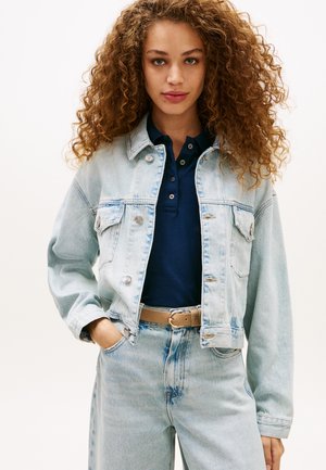 Γυναίκα με κατσαρά μαλλιά που φοράει ανοιχτό μπλε τζιν μπουφάν και τζιν παντελόνι, μπλε πουκάμισο πόλο και μπεζ ζώνη, στέκεται με το ένα χέρι στην τσέπη.