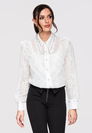 Mujer que lleva una blusa blanca transparente de encaje con cuello adornado y volantes, combinada con pantalones negros de talle alto con cremallera frontal.