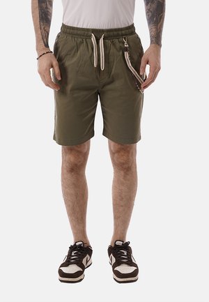 Mann trägt olivgrüne Shorts mit weißem Kordelzug und Kettenaccessoire, kombiniert mit schwarz-weißen Sneakern, stehend vor weißem Hintergrund.