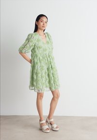 Robe en dentelle verte avec motif floral, décolleté en V, manches bouffantes et taille froncée. Associée à des sandales à talons blanches.