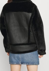 Kvinna iklädd en svart fårskinnsjacka med läderpaneler, tillsammans med en ljusblå denim minikjol, vänd bort.