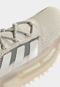 Vaaleanbeige urheilukenkä, jossa on teksturoitu verkko-osa, harmaat raidat ja läpinäkyvä pohja. Varustettu joustavilla nauhoilla ja näkyvällä pehmustuksella.