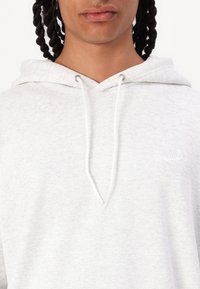 Sudadera gris claro hecha de tejido texturizado, con un bolsillo tipo canguro en la parte frontal y cordones blancos. Incluye un pequeño logo blanco en el pecho.