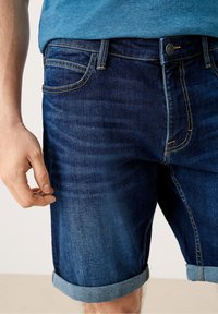 Tummansiniset farkkuShortsit, joissa on hieman haalistunut pinta. Niissä on käännetyt lahkeet ja erottuva keltainen tikkaus. Etupuolella näkyy käsitasku.