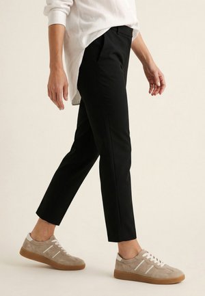 Person trägt schwarze Slim-Fit-Hose, beige Sneakers und ein weißes Langarmshirt, geht vor neutralem Hintergrund.