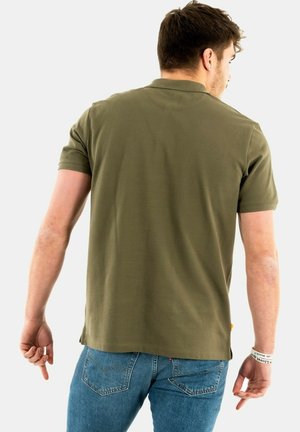 Polo shirt verde oliva con maniche corte e colletto, realizzato in un tessuto liscio. Indossato con jeans blu, che mostrano un taglio casual.