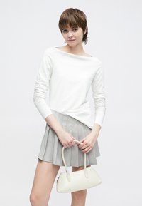 Hvit langermet topp med vid halslinning, kombinert med en grå plissert miniskjørt. Modellen holder en liten kremfarget håndveske med buede håndtak.