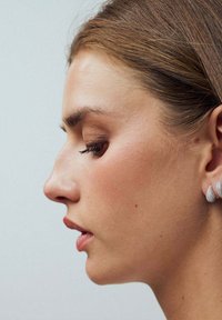 Silverfärgade droppörhängen har en texturerad yta. Modellen har håret uppsatt, vilket framhäver hennes profil och örhängens design.