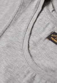 Szary t-shirt z tkaniny z okrągłym dekoltem i widocznymi szwami. Metka z napisem "SUPER PREMIUM" w żółtym kolorze na ciemnym tle.
