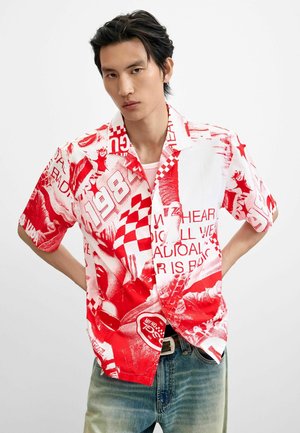 Junger Mann mit kurzen schwarzen Haaren, der ein rotes und weißes grafisches Kurzarmshirt und blaue Jeans trägt, mit den Händen hinter dem Rücken.