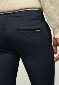 Marinblå byxor med en slät textur, med en bakficka som har knäppning med knapp och en liten märkesetikett. En kraftigt stickad tröja synlig ovanför.