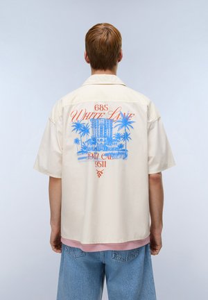 Osoba o krótkich rudawych włosach nosi kremową koszulę z niebieskim nadrukiem palm i budynków oraz pomarańczowym napisem „White Lake” na plecach.