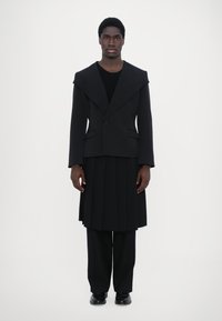 Schwarze maßgeschneiderte Jacke mit breiten Revers, über einem taillierten Oberteil getragen, kombiniert mit einem plissierten schwarzen Rock und maßgeschneiderten Hosen. Glatte Textur insgesamt.