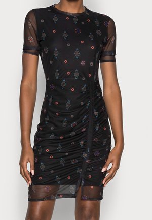 Kvinde iført en tætsiddende sort kjole med gennemsigtige korte ærmer, farverige geometriske og blomsterdekorative mønstre samt rynkning i siden med en frontslids.