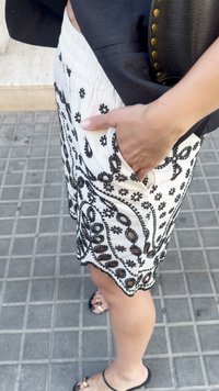 Femeie îmbrăcată în pantaloni scurți albi cu broderie neagră, top negru, purtând o geantă de piele neagră, stând pe un trotuar placat cu țiglă, cu o mână în buzunar.