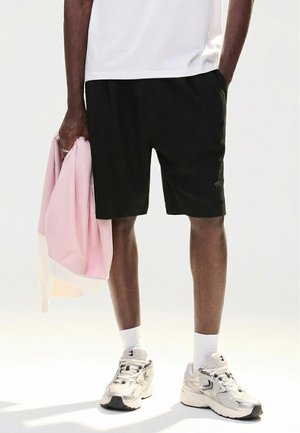 Man draagt zwarte korte broek, witte sokken, zilveren sneakers en houdt een roze jas in zijn linkerhand vast tegen een effen achtergrond.