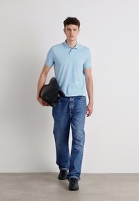 Gaiši zils polo krekls, zili džinsi un melnas kurpes. Modelis tur melnu somu. Vienkāršs dizains, gluda tekstūra un ikdienišķs piegriezums.