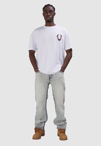 Biała koszulka z krótkim rękawem z czerwoną grafiką, jasnoniebieskie dżinsy i beżowe buty. Krój jest luźny, z widocznymi detalami przeszyć.