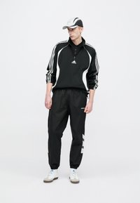 Μαύρο πουλόβερ με φερμουάρ και λευκές ρίγες, μαύρο παντελόνι φόρμας και λευκά αθλητικά παπούτσια. Περιλαμβάνει γκρι και μαύρο καπέλο. Μινιμαλιστικός σχεδιασμός και υλικά.