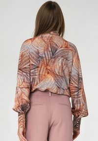 Kobieta nosząca bluzkę z długim rękawem w abstrakcyjne pomarańczowe i brązowe wzory liści, połączoną z spodniami w kolorze mauve o wysokim stanie i dopasowanym kroju.