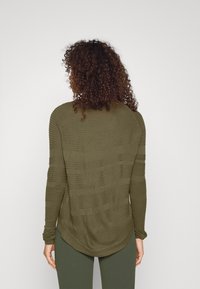 Langärmeliger Pullover in Olivgrün, strukturierter Stoff mit horizontalen Linien, geschwungener Saum und enganliegende Ärmel, kombiniert mit dunkelgrünen Hosen.