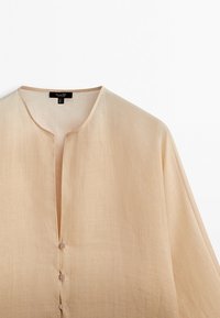Chemise en lin beige clair avec col rond et trois petits boutons sur le devant sur fond blanc.