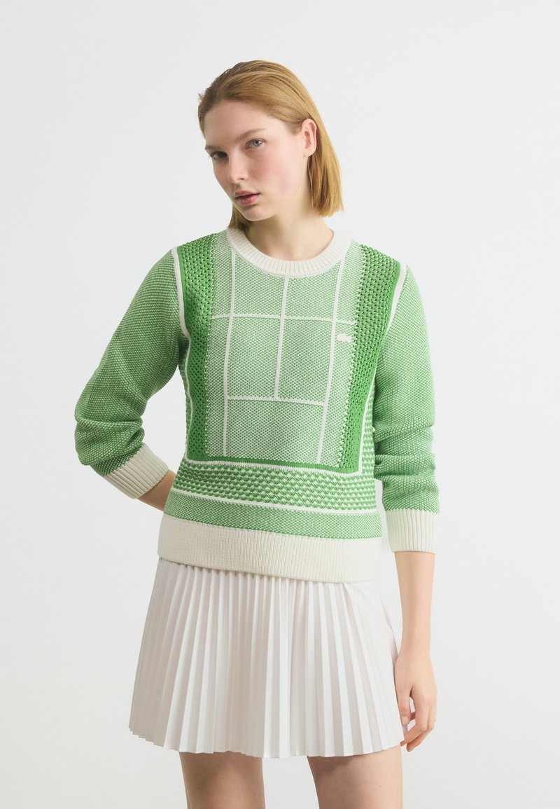 Femme portant un pull en maille verte texturée avec des motifs géométriques blancs et une jupe plissée blanche, debout devant un fond uni.