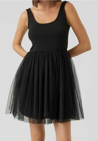 Schwarzes Kleid mit einem eng anliegenden, ärmellosen Oberteil und einem mehrlagigen, durchscheinenden Tüllrock. Glatter Stoff oben, betont durch eine strukturierte Überlagerung.