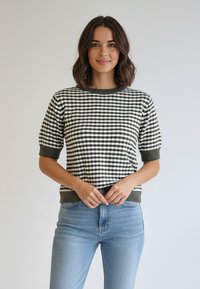 Krótki sweter w zielono-białą kratę z prążkowanym dekoltem i mankietami, zestawiony z jasnoniebieskimi dżinsami. Gładka dzianina.