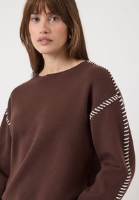 Femme aux cheveux bruns portant un pull marron avec des détails brodés blancs le long des épaules et des manches, sur un fond uni.