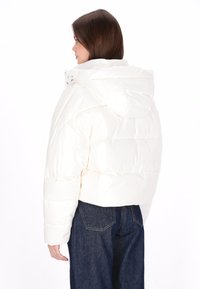 Vatteret jakke i skinnende hvidt materiale, kort design med hætte, quiltet tekstur, båret med mørke denimjeans.